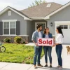 We buy houses for cash, happy couple holding sold sign in front of their home.
